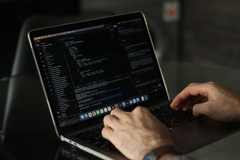 A close-up view of a person's hands typing on a MacBook laptop placed on a dark, reflective table. The laptop screen displays lines of code with a dark background and colorful syntax highlighting, ind